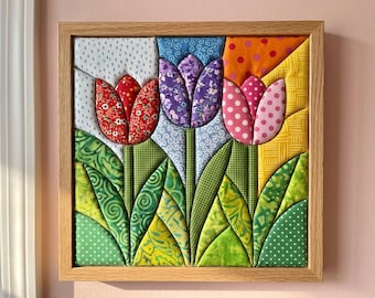 May include: A square fabric wall art piece in a light wood frame featuring three-dimensional quilted tulips. The flowers are red, purple, and pink with various patterns, set against a background of blue, orange, and yellow fabric patches.