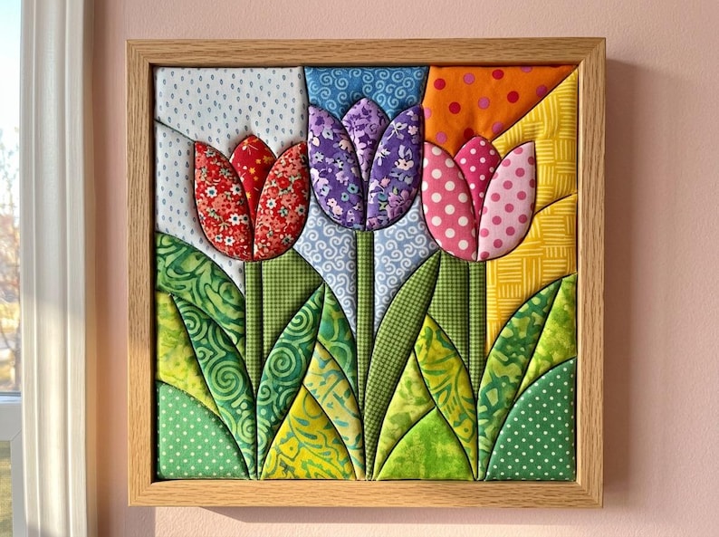 May include: A square fabric wall art piece in a light wood frame featuring three dimensional quilted tulips. The flowers are red, purple, and pink with various patterns, set against a background of blue, orange, and yellow fabric patches.