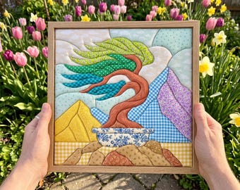May include: A square fabric wall art piece in a light wood frame featuring a quilted bonsai tree with a brown trunk and green, teal, and blue foliage. The tree sits in a blue and white floral pot against a background of yellow, blue, and purple fabric.