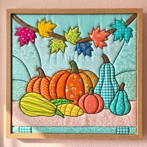 Può includere: Un arazzo quadrato in tessuto in una cornice di legno chiaro che presenta una scena trapuntata di zucche e zucche ornamentali colorate con motivi arancioni, gialli, verdi e blu. Sopra le zucche, un ramo marrone mostra foglie autunnali in tessuto.