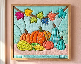May include: A square fabric wall hanging in a light wood frame featuring a quilted scene of colourful pumpkins and gourds in orange, yellow, green, and blue patterns. Above the pumpkins, a brown branch displays various patterned fabric autumn leaves.