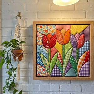 May include: A square fabric wall art piece featuring a quilted tulip design in a light wood frame. The artwork displays three stylized tulips in red, orange, and purple, crafted from various patterned fabrics including gingham, floral, and polka dot prints.