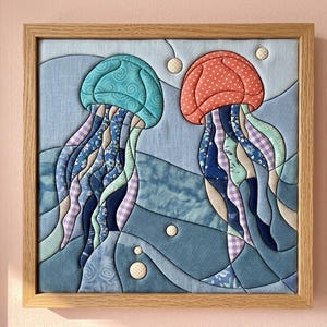 May include: A square fabric wall art piece in a light wood frame featuring a quilted design of two jellyfish. One jellyfish is teal with swirls, the other is coral with white dots. Both have long, flowing tentacles made of various blue patterned fabrics.