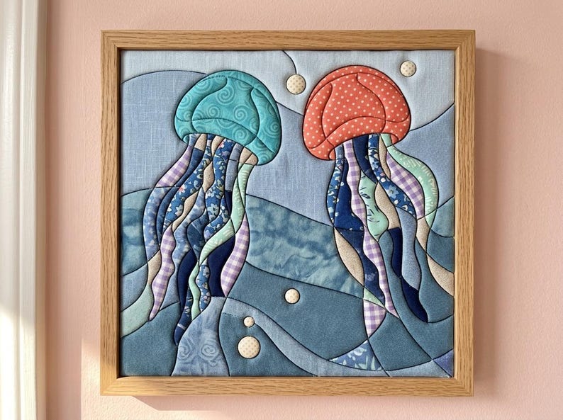 May include: A square fabric wall art piece in a light wood frame featuring a quilted design of two jellyfish. One jellyfish is teal with swirls, the other is coral with white dots. Both have long, flowing tentacles made of various blue patterned fabrics.