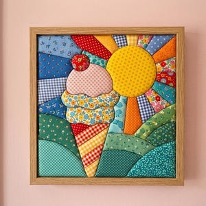 May include: A square fabric wall art piece in a light wood frame featuring a quilted patchwork design of an ice cream cone topped with a cherry, set against a vibrant sunburst background with green rolling hills in various patterned textiles.
