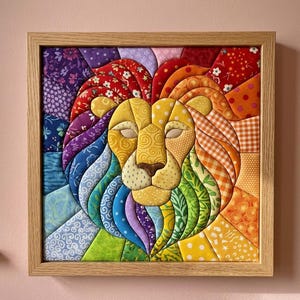 May include: A square fabric wall art piece featuring a stylized lion head constructed from various colorful patterned quilt patches. The lion has a yellow face and a vibrant mane composed of red, orange, yellow, green, blue, and purple fabric segments, all encased in a light-toned wooden frame.
