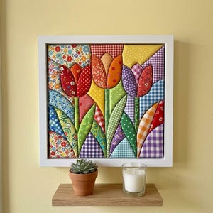 May include: A square wall art piece featuring a fabric-pieced tulip design in a white frame. The tulips are constructed from various colorful patterned fabrics including gingham, floral, and polka dot prints in red, orange, yellow, and purple.
