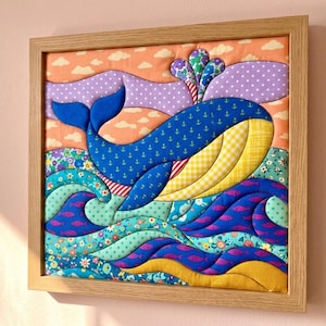Puede incluir: Un cuadro de tela enmarcado que muestra una ballena azul con estampado de anclas, vientre a cuadros amarillos y una escena de agua floral. El fondo incluye nubes naranjas y colinas moradas, todo hecho de parches textiles acolchados.