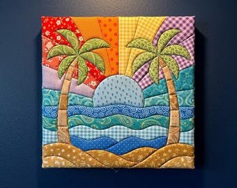 May include: A square fabric wall art piece featuring a quilted tropical scene with two palm trees, a blue sun, and layered waves. The background displays a sunburst pattern in red, orange, yellow, and purple fabrics with various floral and geometric prints.