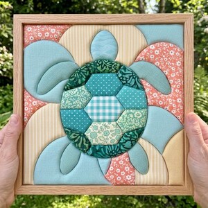 Puede incluir: Una pieza de arte textil enmarcada de forma cuadrada con un motivo de tortuga marina construido a partir de varios parches de tela acolchada. El caparazón está compuesto por patrones hexagonales verdes, mientras que las aletas y el fondo utilizan segmentos de tela azul claro, floral color melocotón y rayas crema.