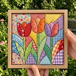 May include: A square fabric wall art piece featuring a patchwork design of three tulips in red, orange, and purple. The flowers are constructed from various patterned textiles including gingham, floral, and polka dot prints, all encased in a light wood frame.