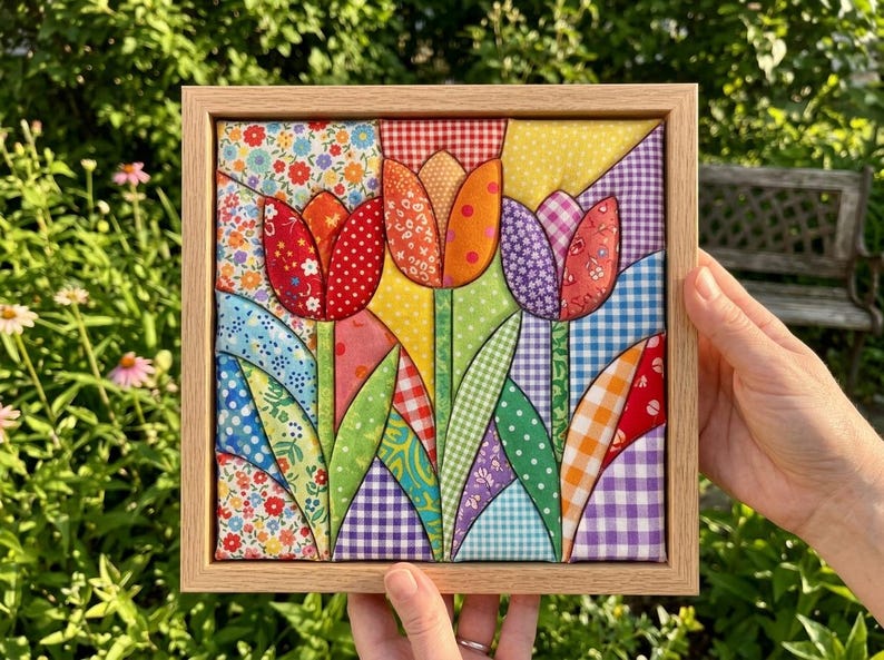 May include: A square fabric wall art piece featuring a patchwork design of three tulips in red, orange, and purple. The flowers are constructed from various patterned textiles including gingham, floral, and polka dot prints, all encased in a light wood frame.