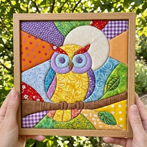May include: A square framed fabric art piece featuring a colourful quilted owl perched on a brown branch. The owl has a yellow body, purple eyes, and red ear tufts, set against a background of geometric fabric patches in orange, blue, and green.