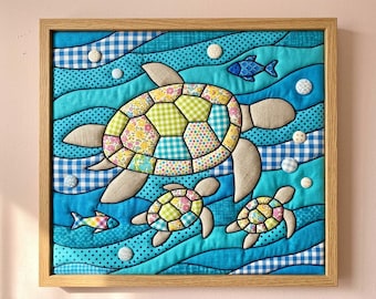 May include: A framed fabric wall art piece featuring a large sea turtle and two smaller turtles swimming through blue waves. The turtles have shells made of colourful patterned fabric patches. Small fabric-covered buttons represent bubbles.
