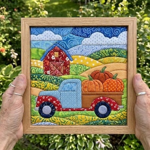 May include: A square fabric quilt art piece in a light wood frame depicting a rural landscape with a red barn, rolling green and yellow hills, white clouds, and a blue pickup truck carrying three orange pumpkins in its cargo bed.