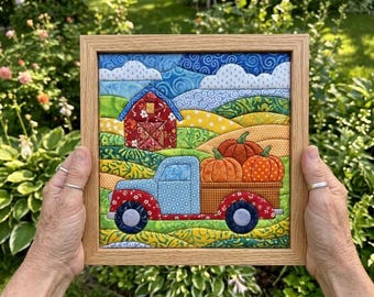 May include: A square fabric quilt art piece in a light wood frame depicting a rural landscape with a red barn, rolling green and yellow hills, white clouds, and a blue pickup truck carrying three orange pumpkins in its cargo bed.