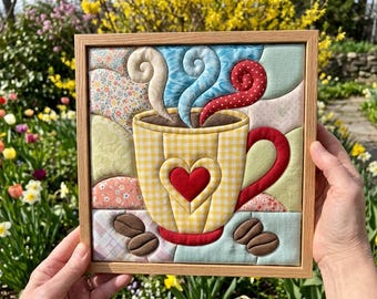 May include: A framed fabric wall art piece featuring a yellow gingham coffee mug with a red heart appliqué. The mug has swirling steam in white, blue, and red polka dot fabric above it, with brown coffee bean appliqués at the bottom corners.