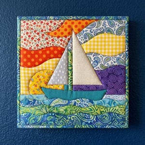 Puede incluir: Un tapiz cuadrado de tela con una escena de velero acolchado. El diseño utiliza varios textiles estampados en naranja, amarillo, morado, verde y azul, representando un barco sobre el agua con un fondo de sol y cielo, todo con un borde azul.