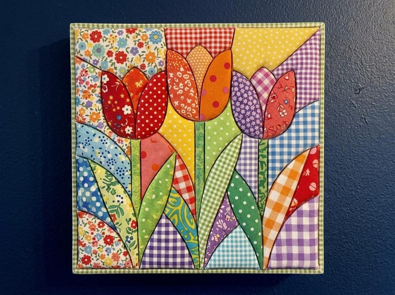 May include: A square fabric wall hanging featuring a pieced quilt design of three tulips. The flowers and leaves are constructed from various cotton fabrics in red, orange, yellow, purple, and green, showcasing floral, gingham, and polka dot patterns.