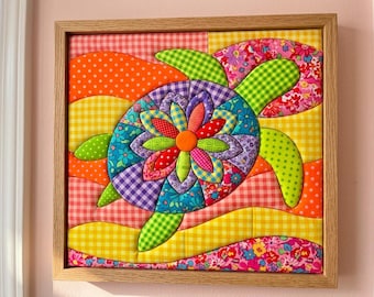 May include: A square framed fabric wall art piece featuring a quilted sea turtle design. The turtle shell is composed of colourful floral, gingham, and polka dot fabric petals. The background consists of wavy fabric strips in yellow and orange.