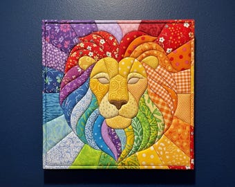 May include: A square fabric wall hanging featuring a stylised lion head constructed from various colourful patterned quilt pieces. The lion's mane radiates outwards in segments of red, orange, yellow, green, blue, and purple fabric prints.