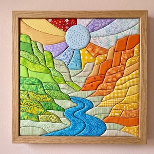 Op de afbeelding: Een ingelijst stoffen wandkunstwerk met een landschapsscène met een blauwe rivier die stroomt tussen groene heuvels aan de linkerkant en oranje heuvels aan de rechterkant. Een ronde zon met een blauw patroon straalt kleurrijke stoffen stralen uit.