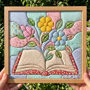 Peut inclure: Une œuvre d'art murale en tissu encadrée de forme carrée présentant un motif matelassé d'un livre ouvert avec des fleurs en pleine floraison. Le livre a des pages beiges à motifs, tandis que les fleurs sont jaunes, bleues et roses.
