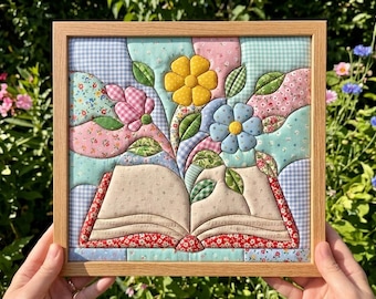 May include: A square framed fabric wall art piece featuring a quilted design of an open book with blooming flowers. The book has beige pages with a subtle pattern, while the flowers are yellow, blue, and pink, surrounded by green fabric leaves.