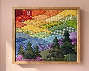 May include: A framed fabric wall art piece featuring a landscape of rolling hills and evergreen trees. The scene is constructed from various patterned and solid-coloured fabric scraps in shades of red, orange, yellow, green, blue, and purple.