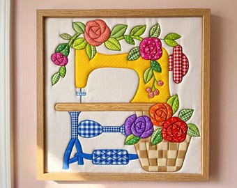 May include: A framed fabric wall art piece featuring a yellow sewing machine appliqué on a white background. The machine is decorated with colourful fabric roses, green leaves, and a woven basket. The base has blue and white checkered patterns.