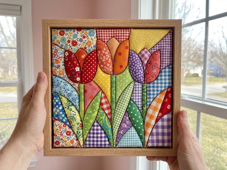 May include: A square fabric wall art piece featuring a quilted tulip design in a light wood frame. The artwork displays three stylized tulips made from various colorful patterned fabrics, including floral, gingham, polka dot, and plaid prints.