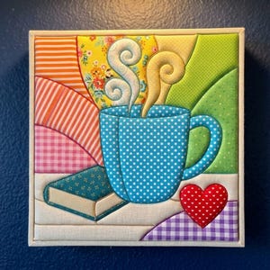 May include: A square fabric wall art piece featuring a blue polka-dot mug with swirling steam, a small green book, and a red heart with white dots. The background consists of colorful quilted fabric sections in orange, yellow, and green patterns.