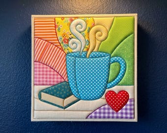 May include: A square fabric wall art piece featuring a blue polka-dot mug with swirling steam, a small green book, and a red heart with white dots. The background consists of colourful quilted fabric sections in orange, yellow, and green patterns.