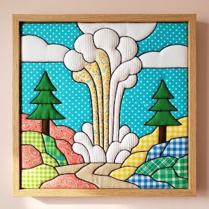 以下が含まれることがあります： 明るい木製のフレームに入った正方形の布製ウォールアートで、キルト風の風景が描かれています。青い水玉模様の空を背景に、2本の緑の松の木の間の小道の上に、中央の白い雲のような噴煙が立ち上る様子が表現されています。