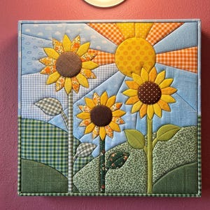 May include: A square fabric wall art piece featuring a quilted sunflower scene. Three sunflowers with brown centres and yellow petals stand in green hills. A yellow sun with orange rays shines in a light blue sky with white clouds and patterns.