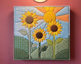 May include: A square fabric wall art piece featuring a quilted sunflower scene. Three sunflowers with brown centres and yellow petals stand in green hills. A yellow sun with orange rays shines in a light blue sky with white clouds and patterns.