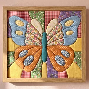 May include: A square framed wall art piece featuring a fabric butterfly made from various quilted patches. The butterfly has blue, orange, and floral patterned wings with a central teal body, all set against a background of red gingham and green floral fabric.