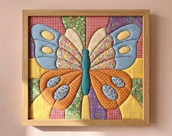 May include: A square framed wall art piece featuring a fabric butterfly made from various quilted patches. The butterfly has blue, orange, and floral patterned wings with a central teal body, all set against a background of red gingham and green floral fabric.
