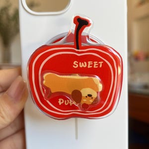 May include: White phone case with a red apple-shaped pop socket. The pop socket has the word "SWEET" and a cartoon dog illustration. The phone case is being held by a person.