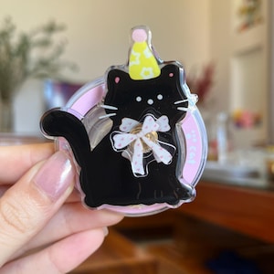 May include: A black cat-shaped acrylic pin with a yellow party hat and a white bow. The pin is set on a pink circle. The cat has white whiskers and is smiling. The pin is held by a hand.