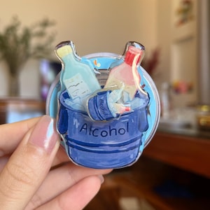 May include: A clear, round sticker featuring a blue bucket filled with two bottles of alcohol. The bucket is labeled "Alcohol" in white text. One bottle is light blue, the other is pink and red.