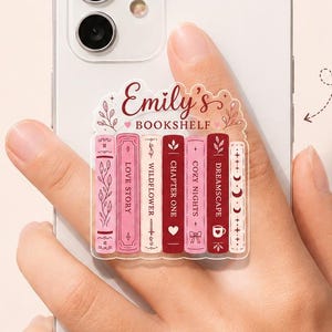 May include: A custom phone grip featuring a bookshelf design with the text Emily's BOOKSHELF and various book titles in shades of pink and red. The accessory is attached to the back of a white smartphone, which also functions as a phone stand.