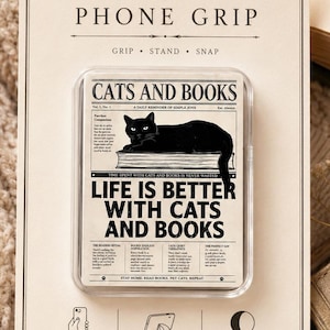 May include: A rectangular phone grip in retail packaging featuring a black cat illustration on a book. The text reads: "CATS AND BOOKS," "LIFE IS BETTER WITH CATS AND BOOKS," and "GRIP, STAND, SNAP." The packaging includes icons for secure grip, built-in stand, and repositionable features.