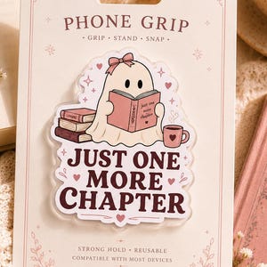 Könnte beinhalten: Ein Handygriff auf einer rechteckigen Trägerkarte mit einer Geisterillustration, die ein Buch liest, mit dem Text "JUST ONE MORE CHAPTER". Der Geist trägt eine rosa Schleife und sitzt neben einem Bücherstapel und einer Tasse. Die Karte enthält den Text "PHONE GRIP", "GRIP • STAND • SNAP", "STRONG HOLD • REUSABLE" und "COMPATIBLE WITH MOST DEVICES".