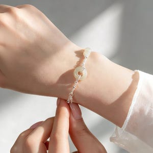 Op de afbeelding: Een delicate zilveren armband met een lichtgroene jade donut-vormige hanger. De armband heeft een delicate ketting met kleine zilveren kralen en een kleine jade kraal. De armband wordt om een pols gedragen.