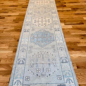 Könnte beinhalten: Ein langer, schmaler Läuferteppich in hellblauen und cremefarbenen Tönen. Das Design umfasst eine Reihe sich wiederholender geometrischer Medaillons und ein Randmuster mit stilisierten ankerartigen Motiven entlang der gesamten Teppichlänge.
