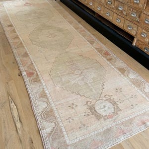 Könnte beinhalten: Ein rechteckiger Teppich im Vintage-Stil mit verblichenem, abgenutztem Aussehen. Der Teppich hat ein zentrales Design in gedämpftem Beige und Olivgrün, umrahmt von einem Rand aus Rosa, Braun und Creme. Der Teppich liegt auf einem Holzboden.