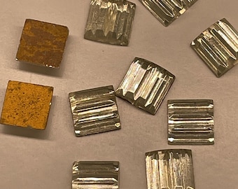 May include: A collection of small square embellishments, including several clear, faceted glass pieces with a ribbed texture and two solid, flat, mustard-yellow square pieces with a textured, matte finish, arranged in a scattered formation.