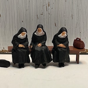 May include: Three miniature figures of nuns wearing black habits and white wimples are sitting on a wooden bench. A brown suitcase is on the bench next to the nuns.