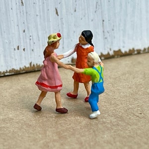 May include: Three miniature figures of children holding hands and dancing in a circle. The girl on the left is wearing a pink dress and a yellow hat with a red flower. The girl in the middle is wearing an orange dress. The boy on the right is wearing a green shirt, blue overalls, and a white hat.